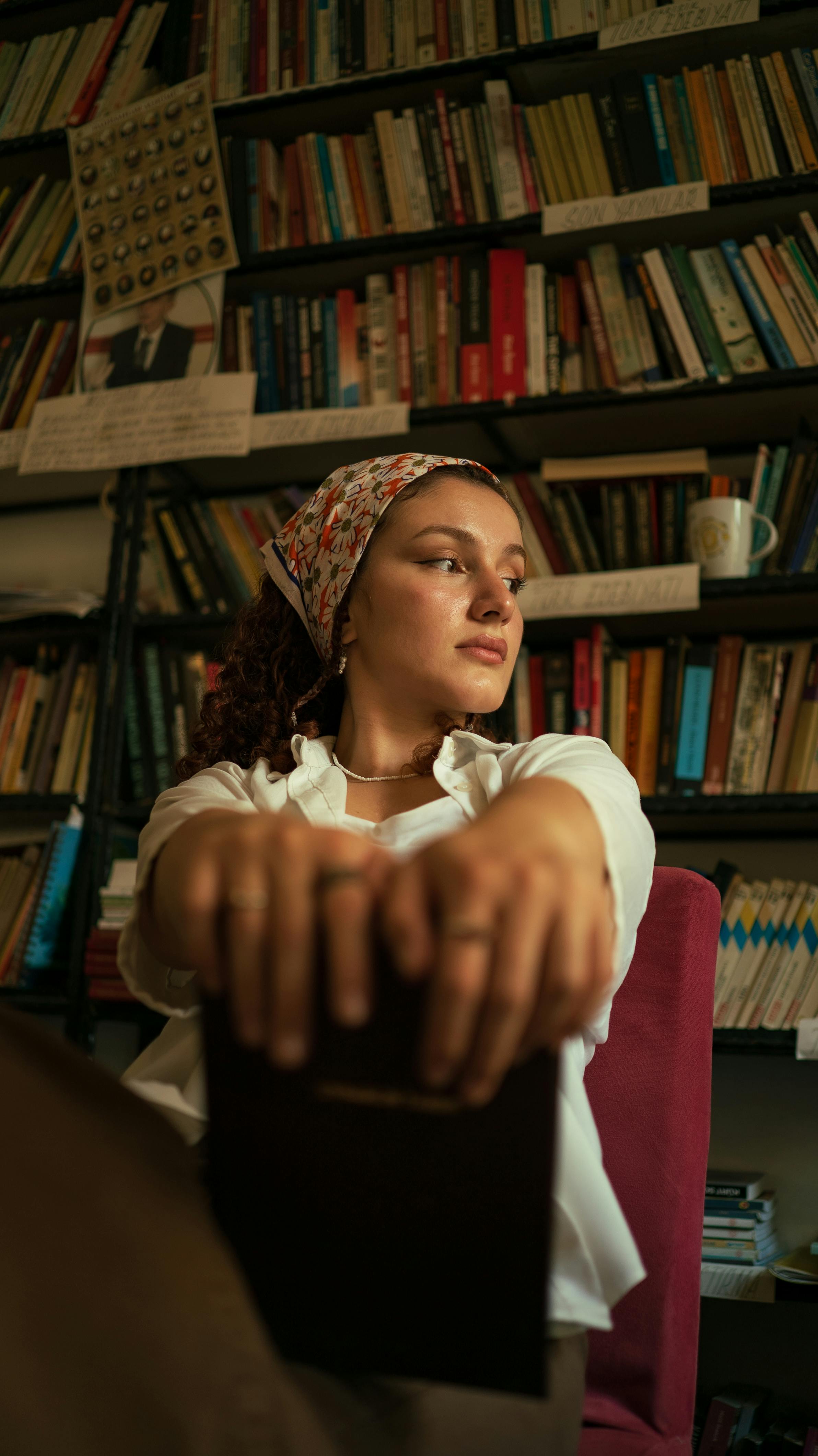 A serene woman sitting in a library, holding a book, surrounded by bookshelves.