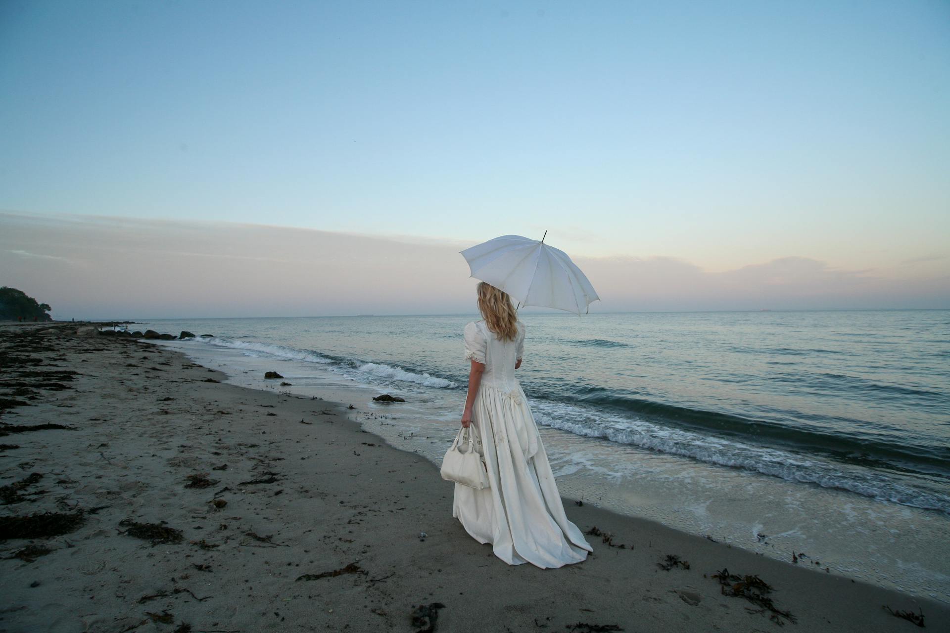 A serene scene of a woman in a white dress holding an umbrella on the beach at twilight.