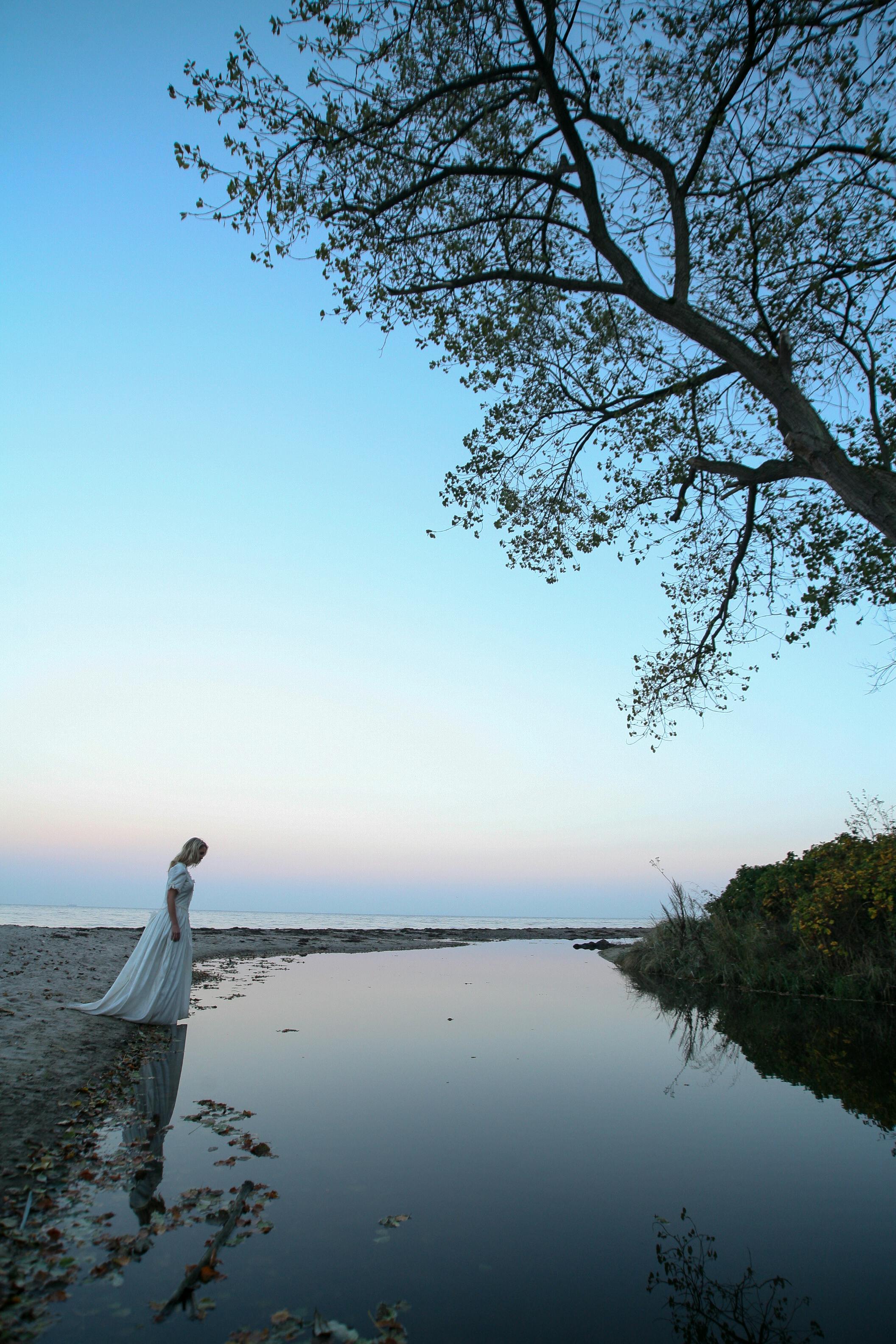 一名身穿白色连衣裙的女子站在湖边，湖面在宁静的天空下反着光，营造出一种平静的感觉