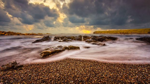 A long exposure shot capturing waves and dramatic clouds at sunset over rocky shores.
