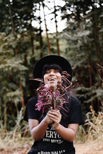 A smiling woman in a hat enjoys holding flowers in a serene forest scene.