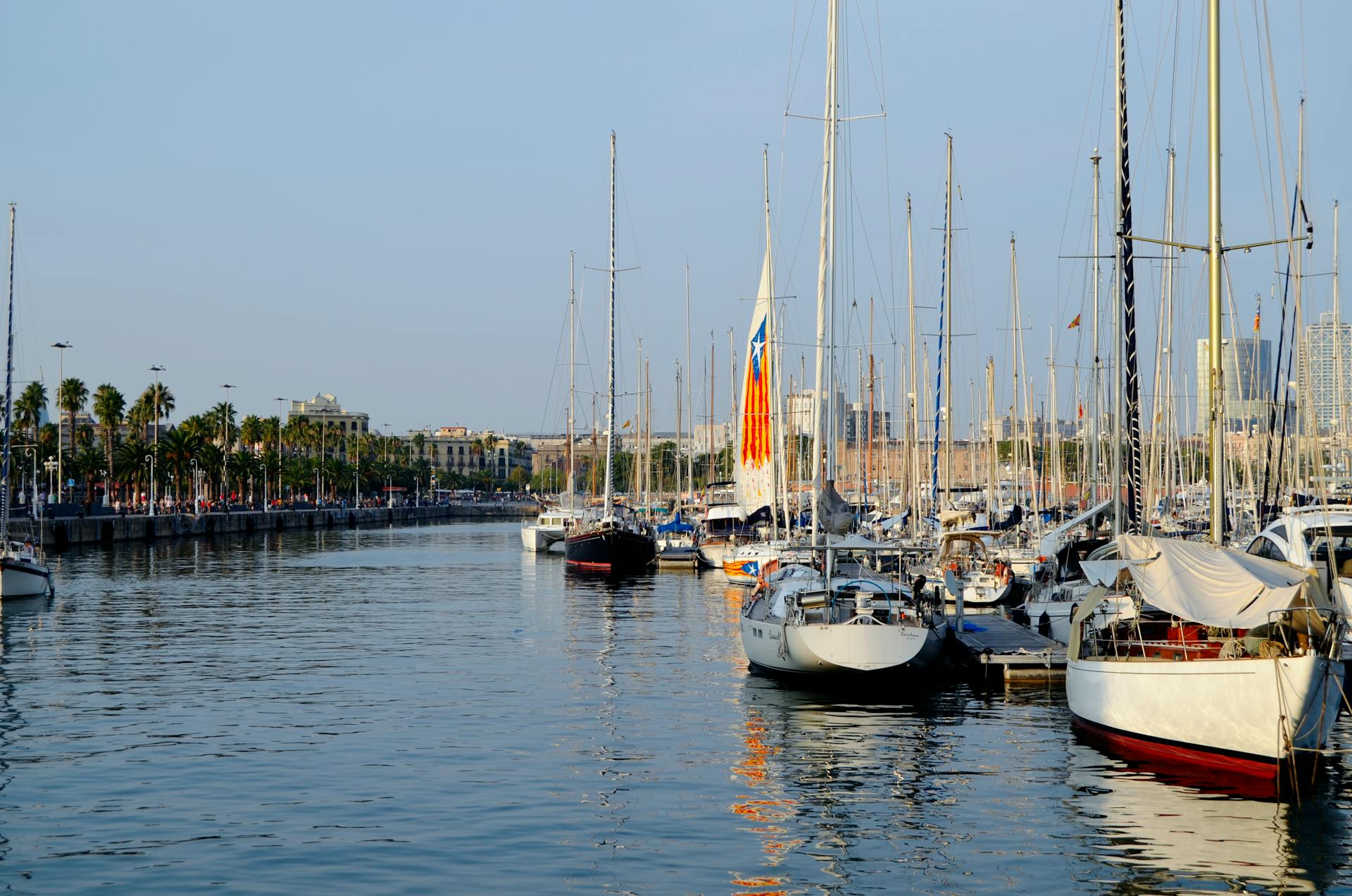 Scenic view of sailboats at Barcelona's Port Vell marina with city skyline in the background.