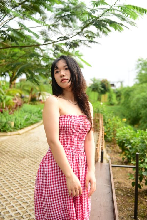 Free Asian woman in a summer park wearing a red gingham dress, standing on a walkway surrounded by lush greenery. Stock Photo