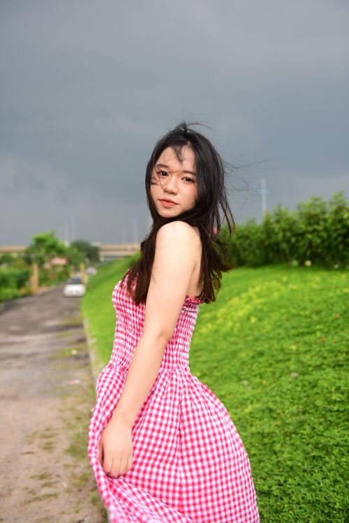 Free Young woman in a red checkered dress poses on a rural road under dark clouds. Stock Photo