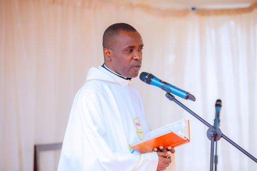 Priest in white robe delivering a sermon with a microphone and holding a Bible.