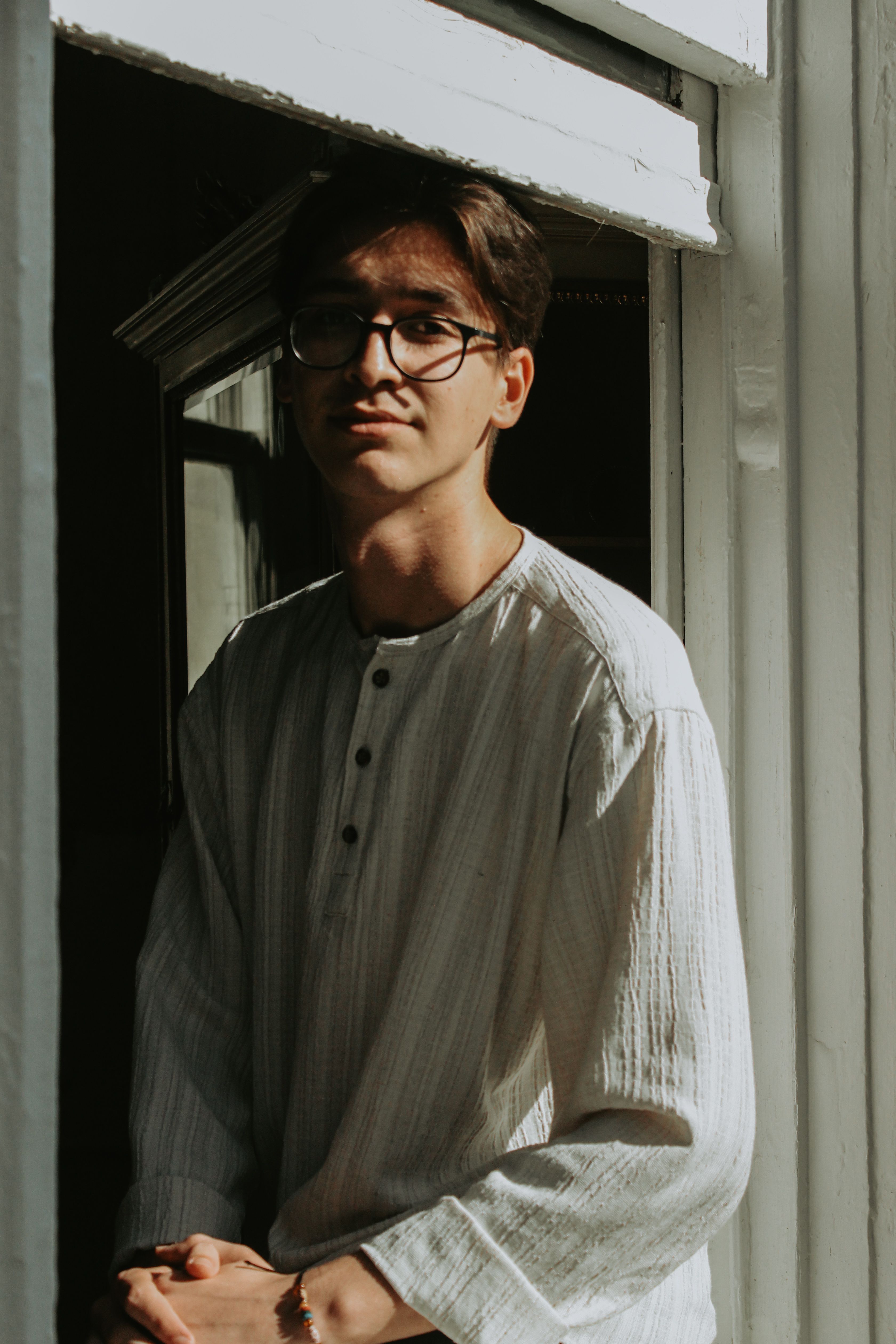 A young man wearing glasses stands by a sunlit window, casting a distinct shadow.