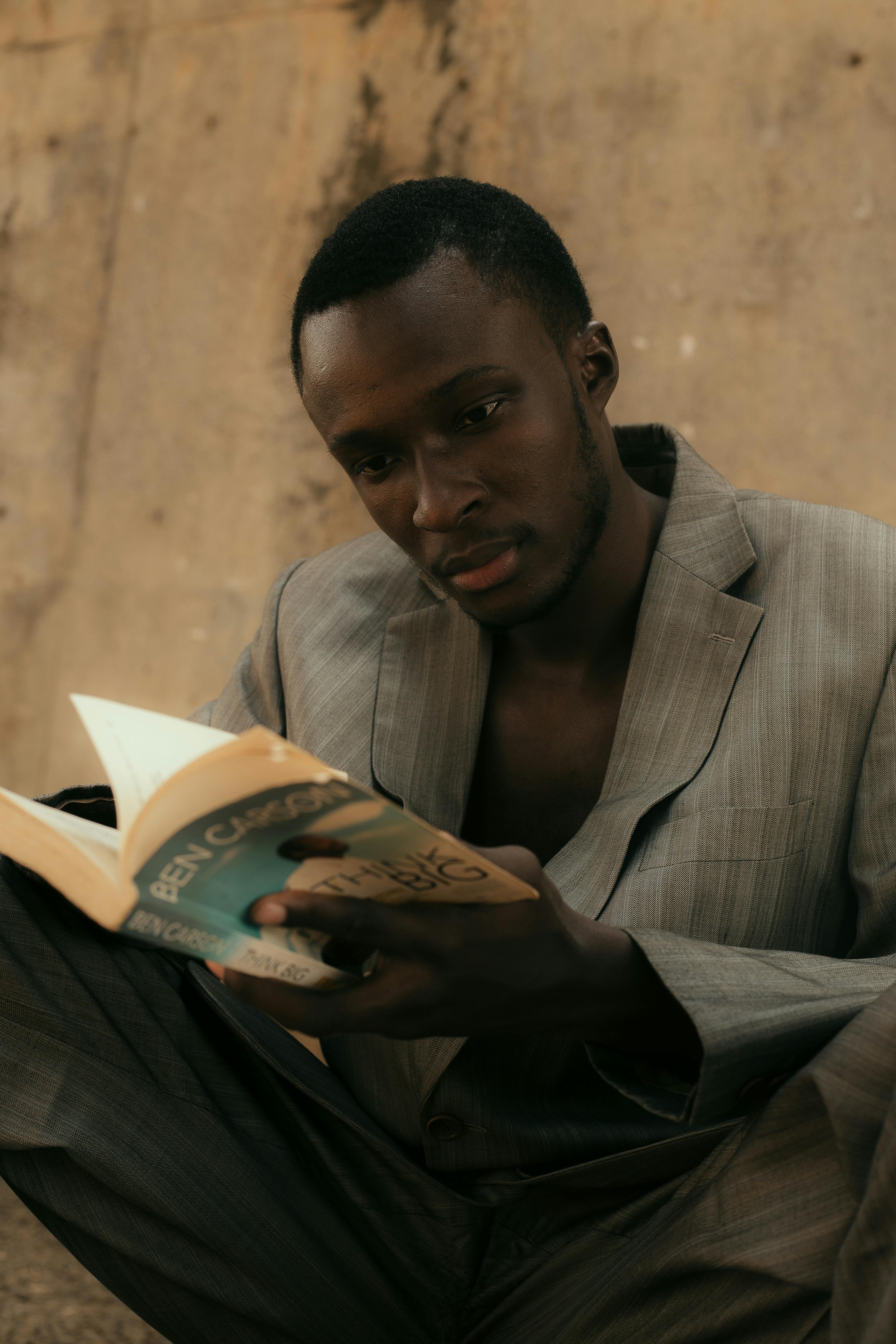 An African American man in a suit reading a book outdoors, embodying concentration.