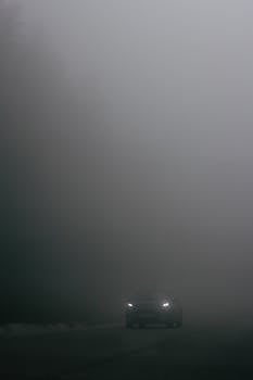 A car's headlights pierce through dense fog on a deserted road, creating a moody and atmospheric scene.