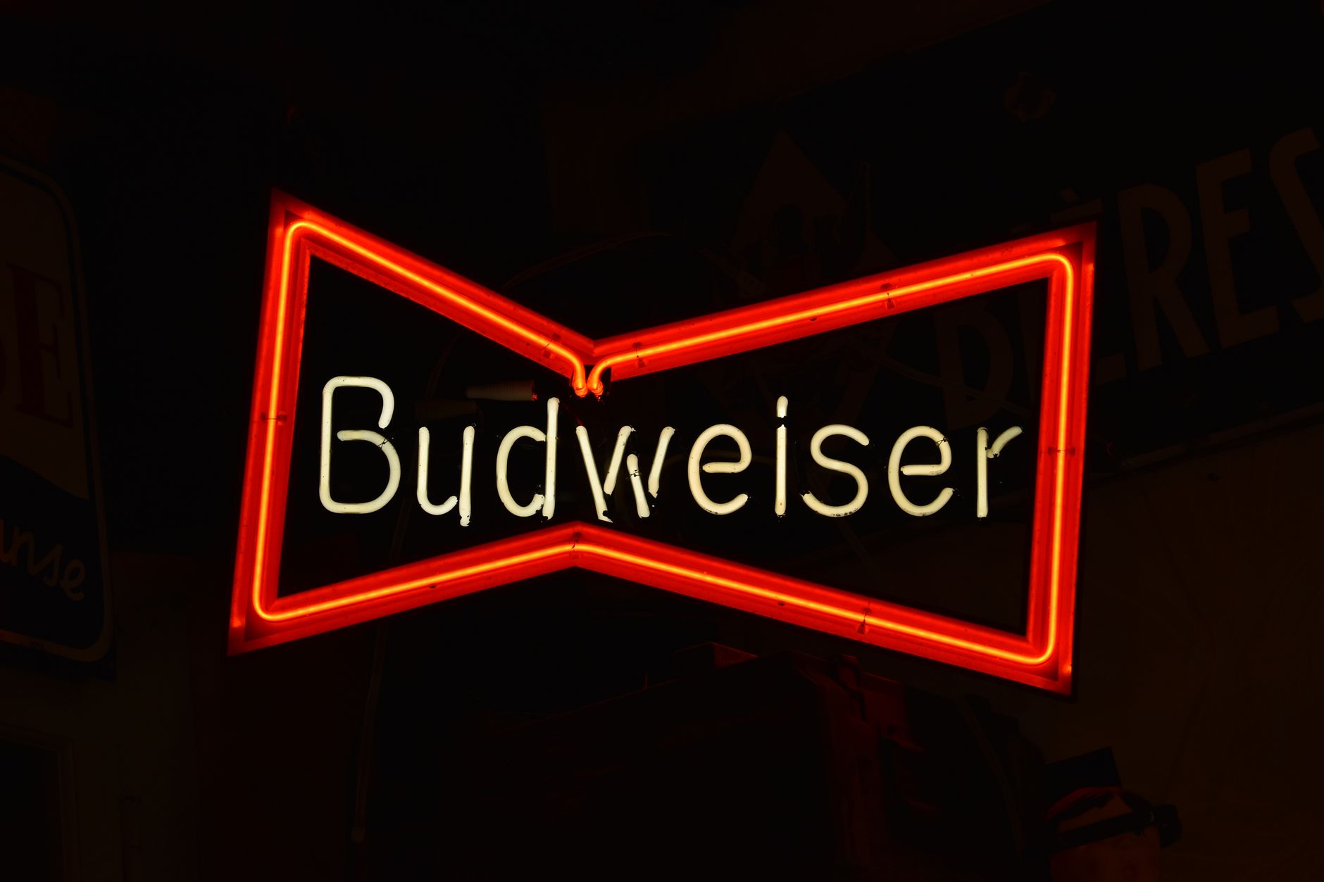A glowing Budweiser neon sign with red and white lighting, showcasing the brand logo against a dark background.