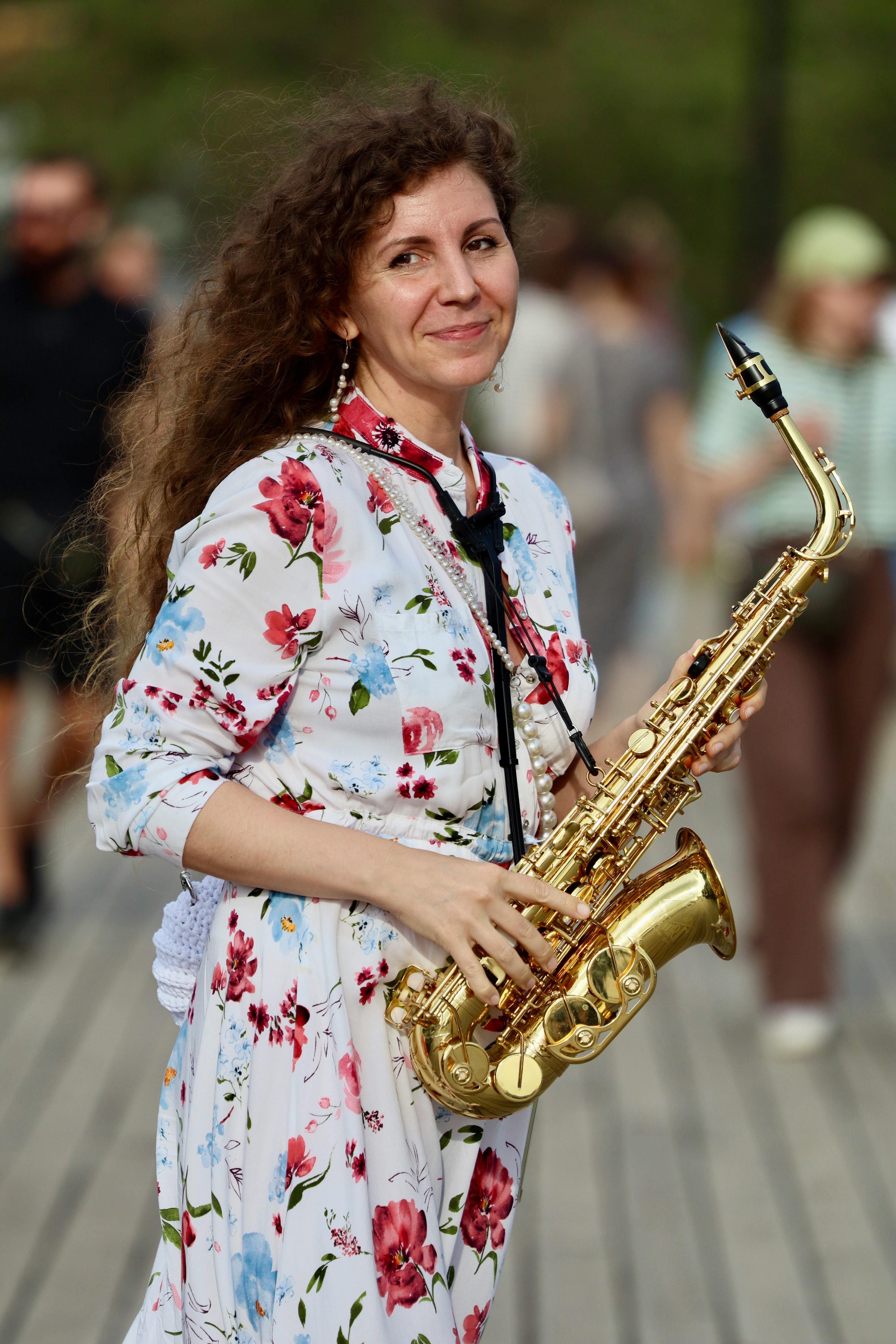 Woman in a floral dress smiling while holding a saxophone outdoors.