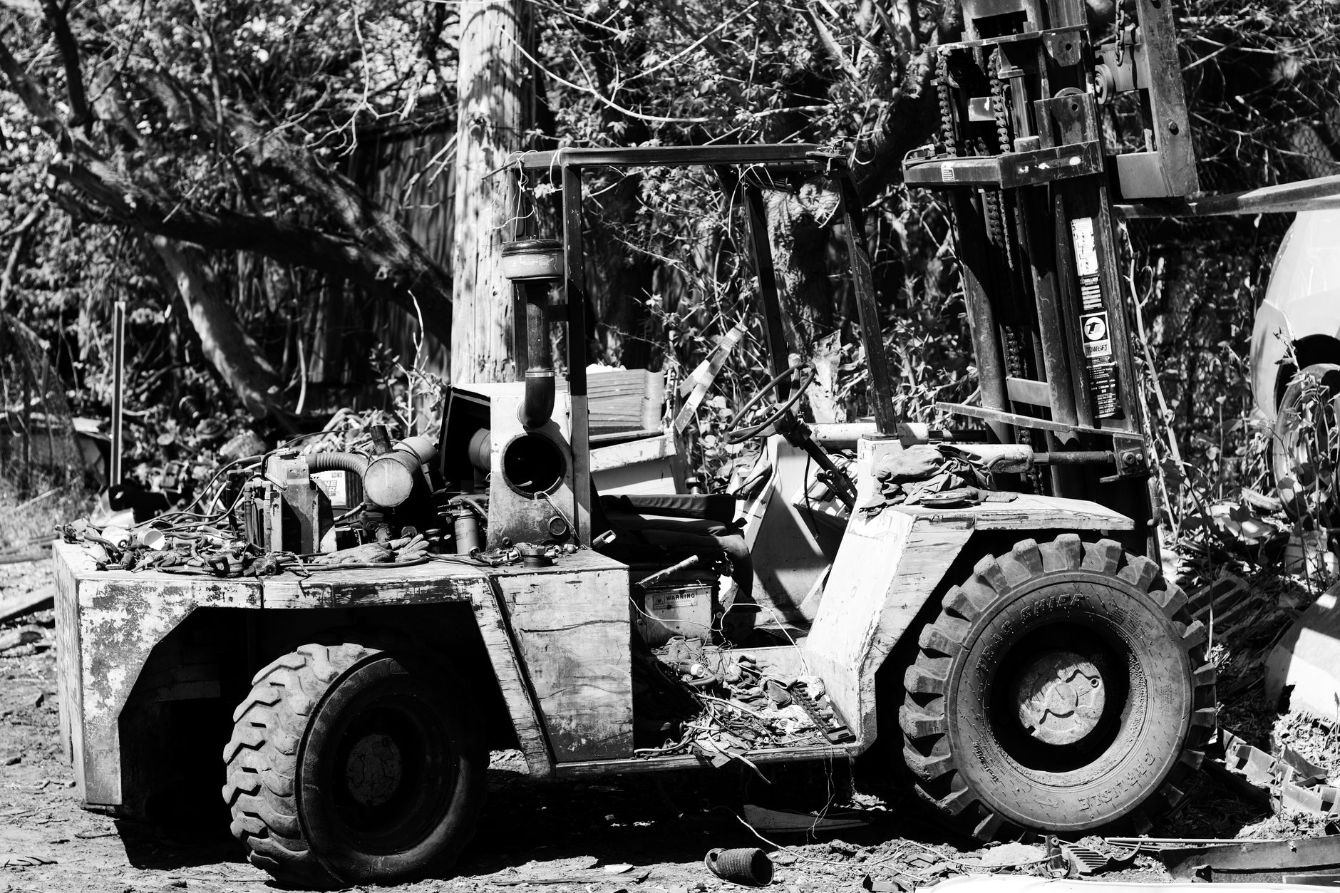 Black and white photo of an abandoned, damaged forklift amidst overgrown vegetation.
