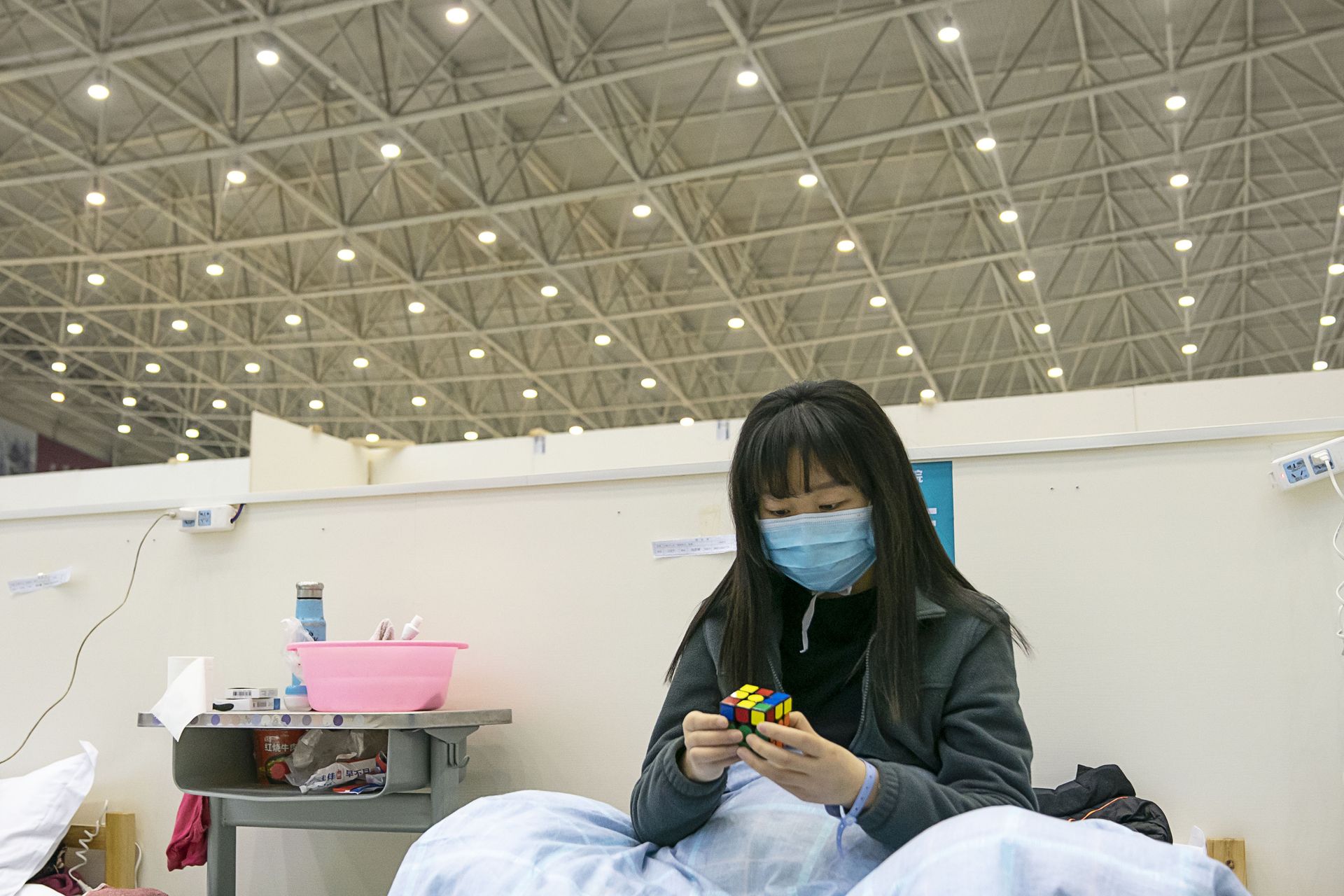 A woman wearing a mask solves a Rubik's Cube in a hospital ward with high ceilings.