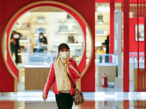 Free A woman in a shopping mall wearing a face mask, scarf, and cap amidst modern red architecture. Stock Photo