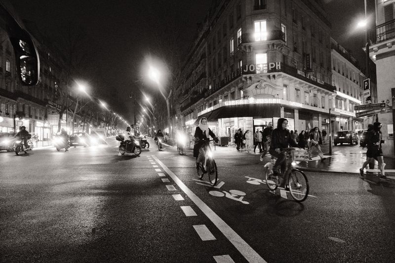 Cyclists and scooters navigate through Parisian streets at night, creating a bustling urban scene.