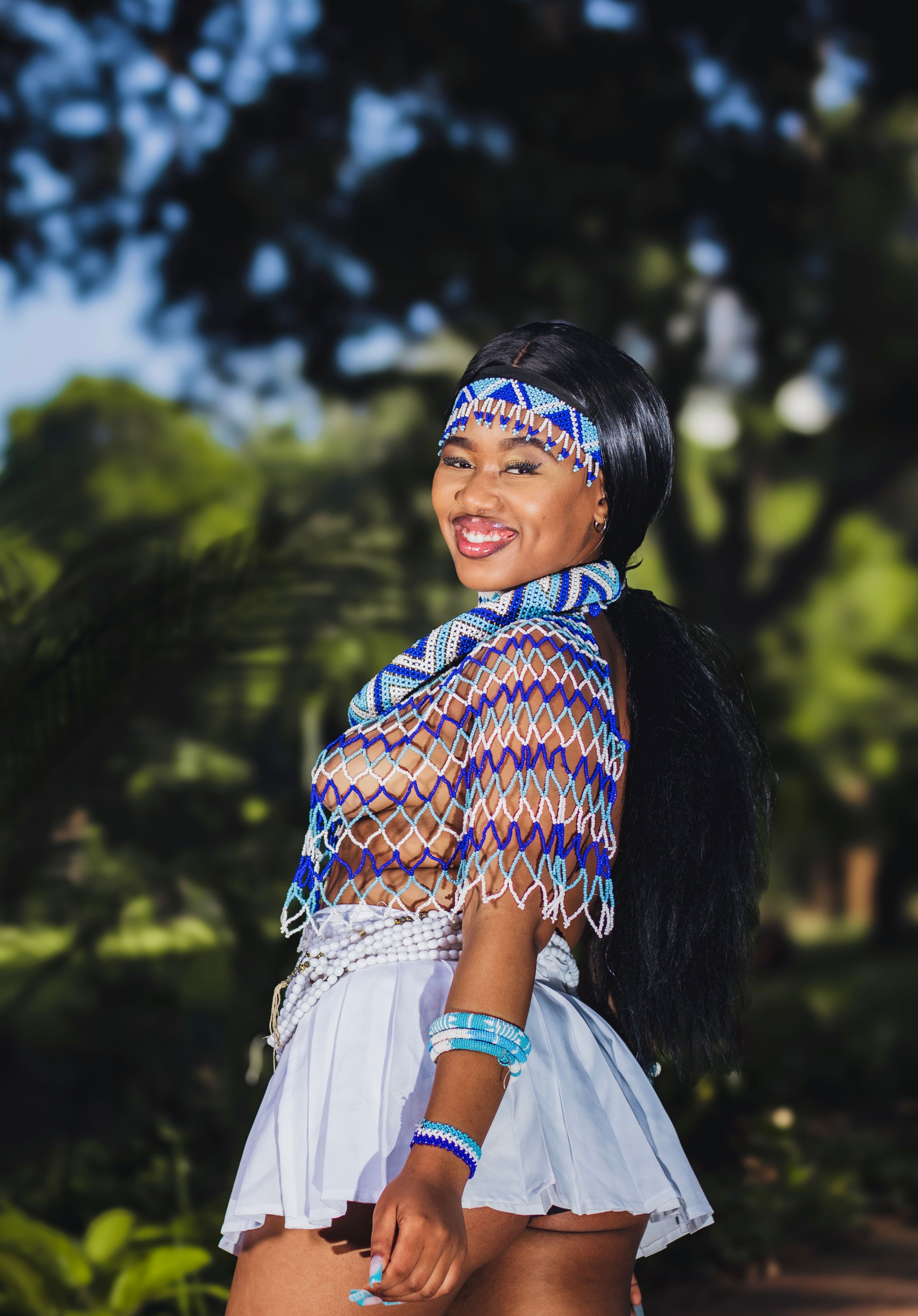 A woman in traditional beaded attire smiles while posing outdoors in a sunny park.