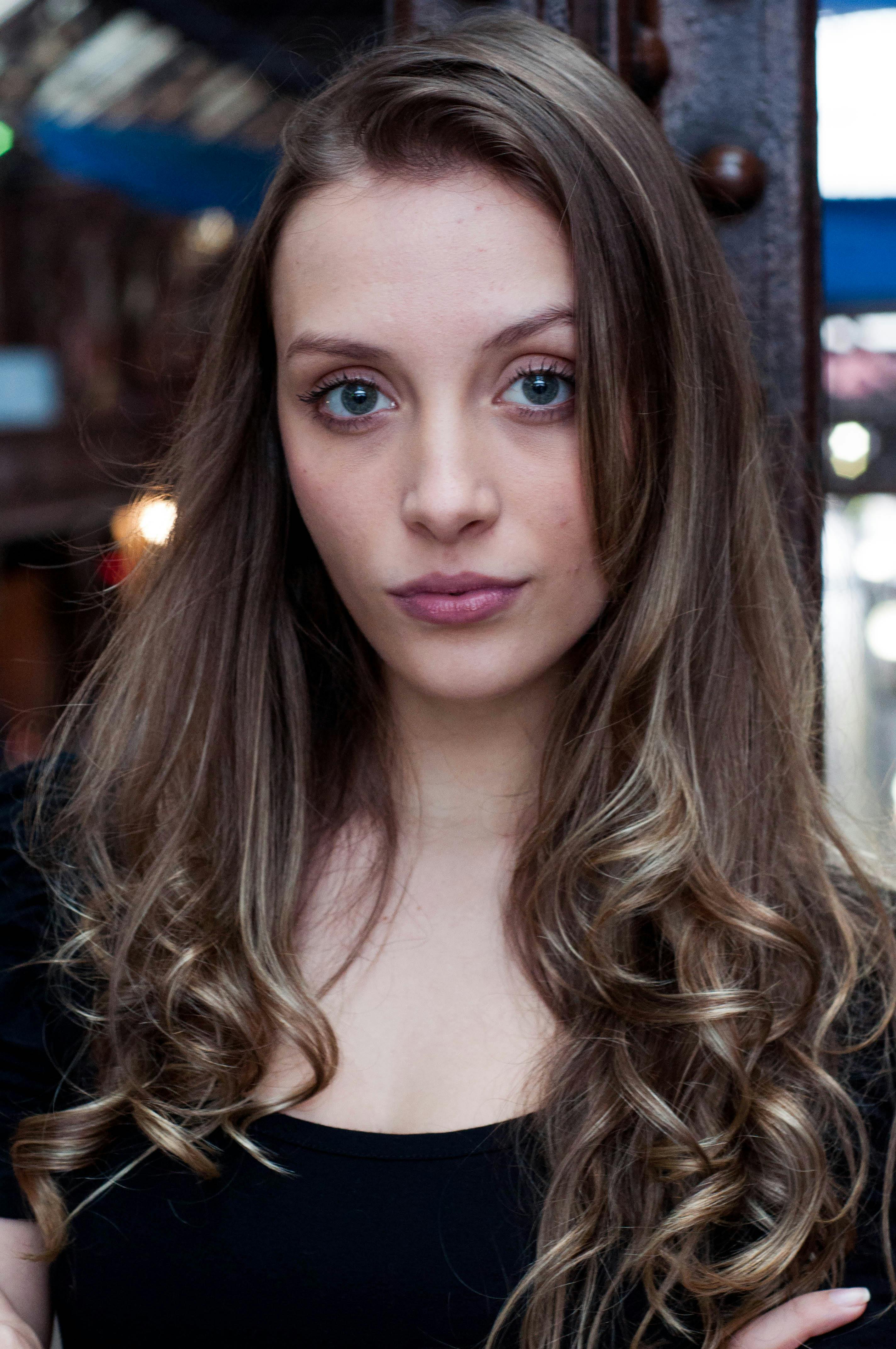 Close-up portrait of a woman with long curly hair and striking eyes, in an urban outdoor setting.