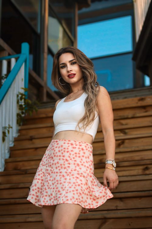 Free Stylish woman in a tank top and floral skirt confidently poses on wooden stairs outdoors. Stock Photo