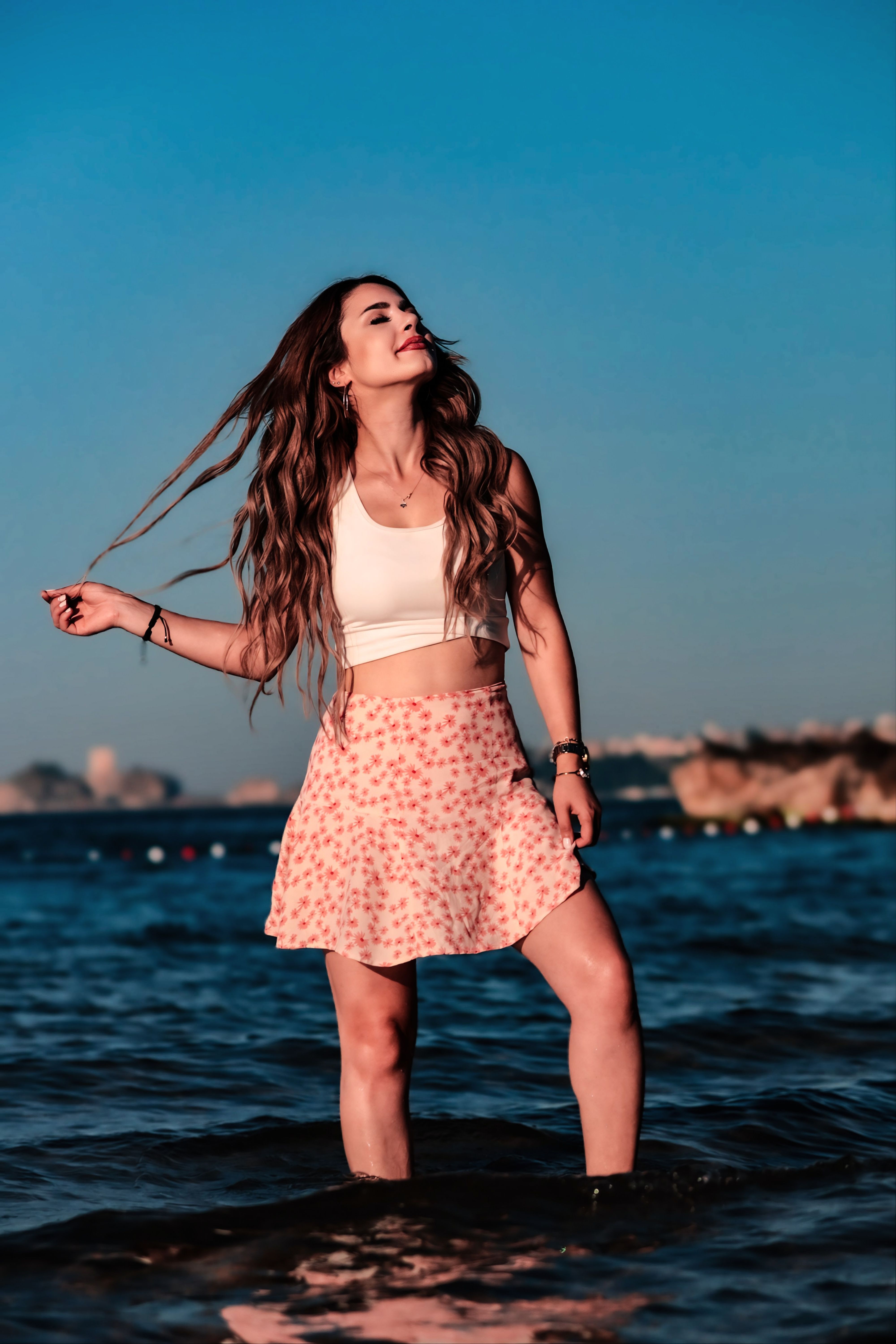 Woman with brown hair standing in the sea, wearing a tank top and skirt, enjoying a sunny day.