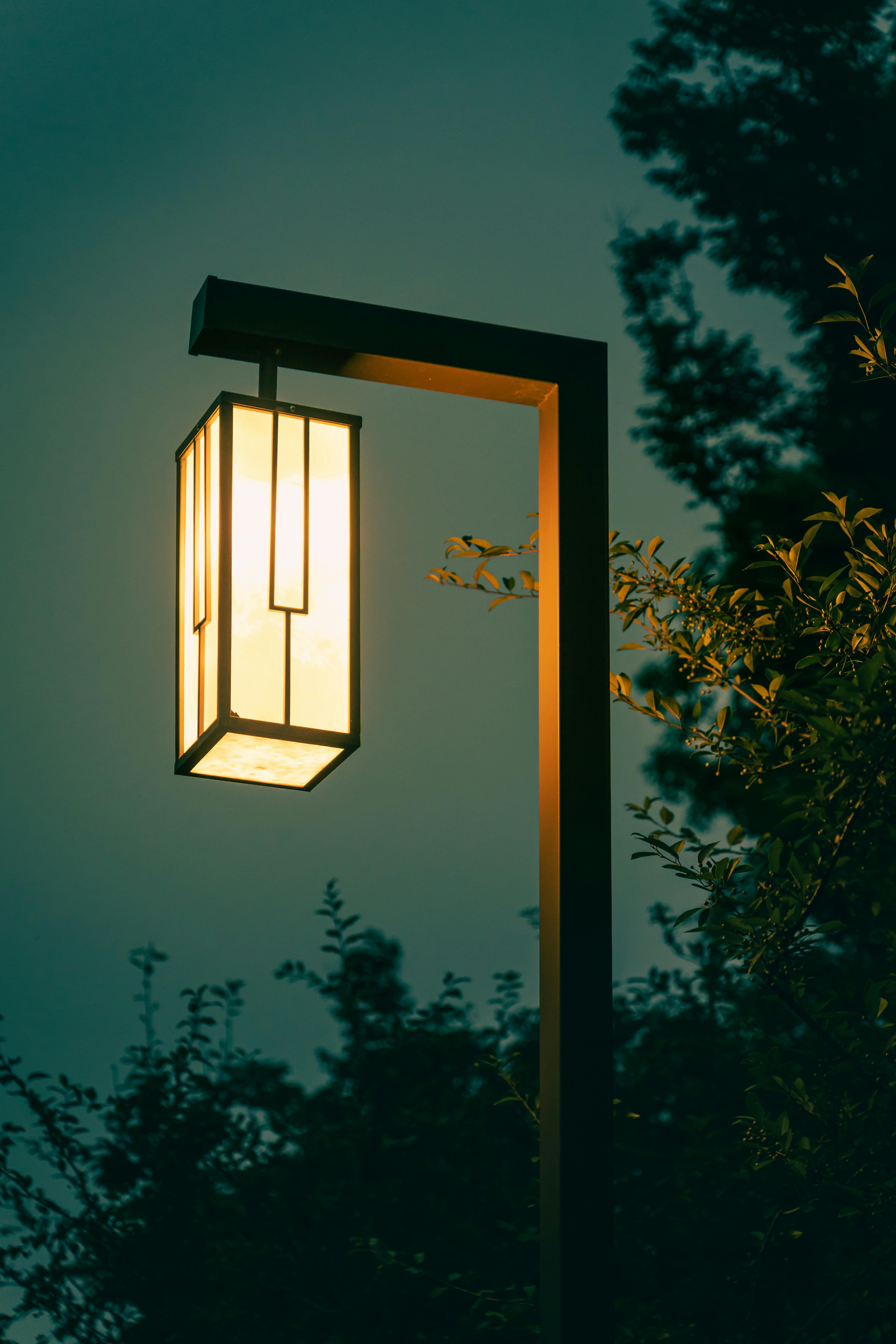 Warm glow of a garden lamp at dusk with trees in the background, creating a peaceful ambiance.