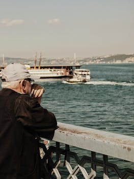 在一个阳光明媚的日子里，一位年长的男子倚着桥栏杆，望着渡轮横渡大海