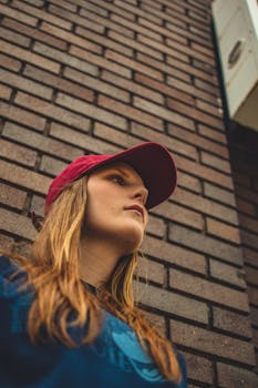 A young woman wearing a red cap stands outdoors against a brick wall, looking away thoughtfully.