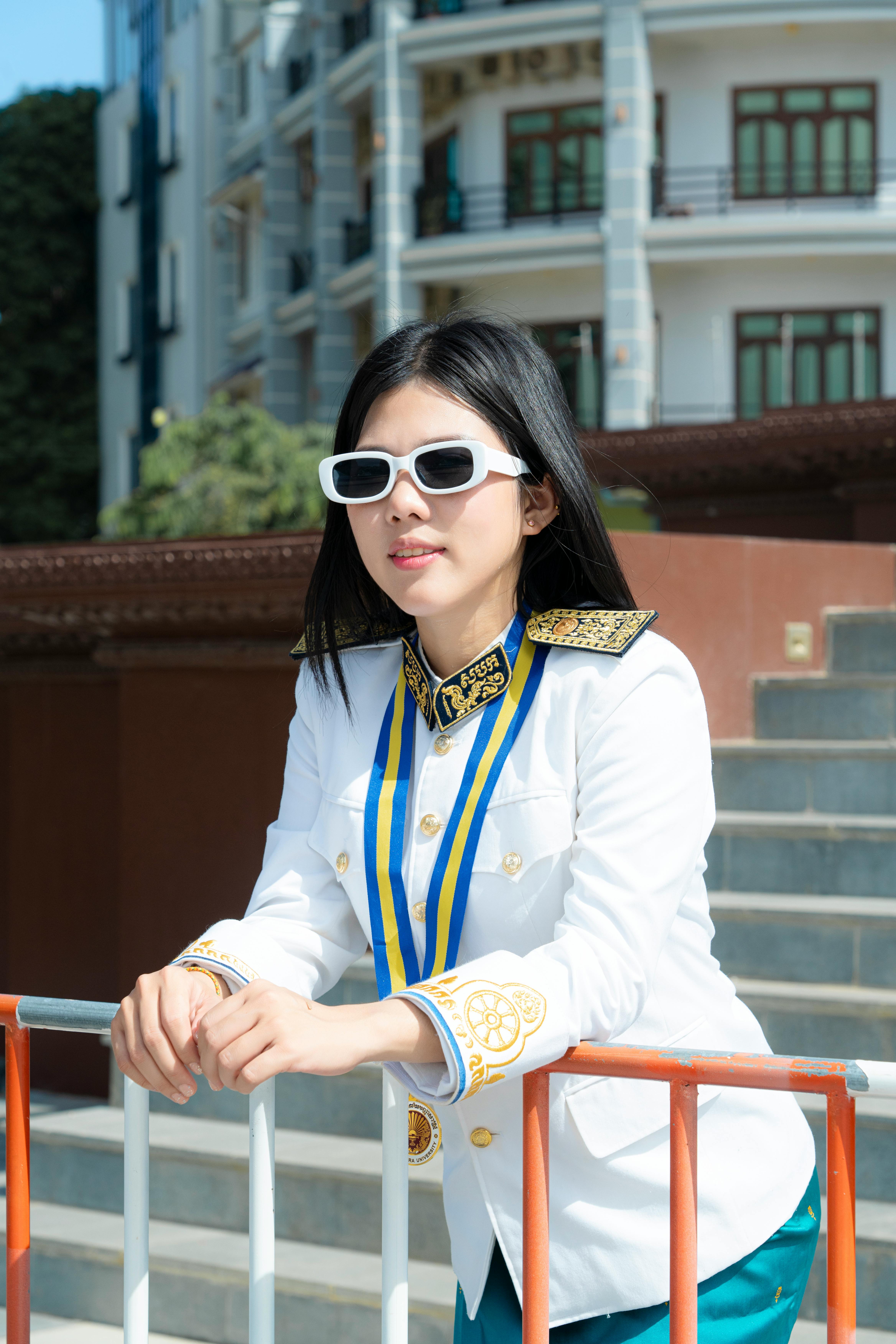 Elegant young woman in white uniform and sunglasses at an outdoor graduation setting.