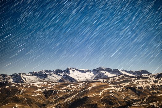 Long exposure shot capturing star trails above a snowy mountain range during night.