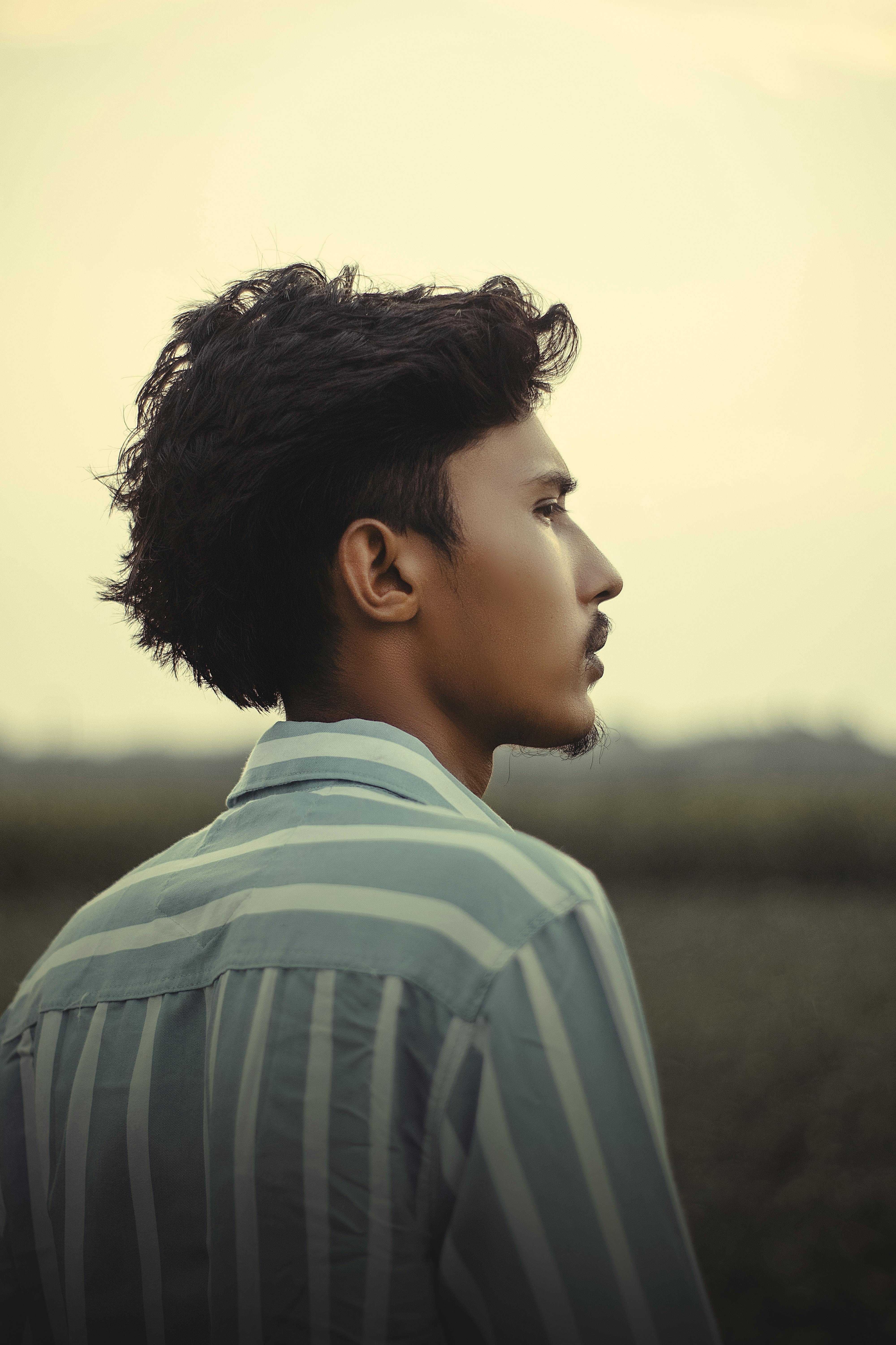 A stylish young man with short hair gazes into the distance in a serene outdoor setting.