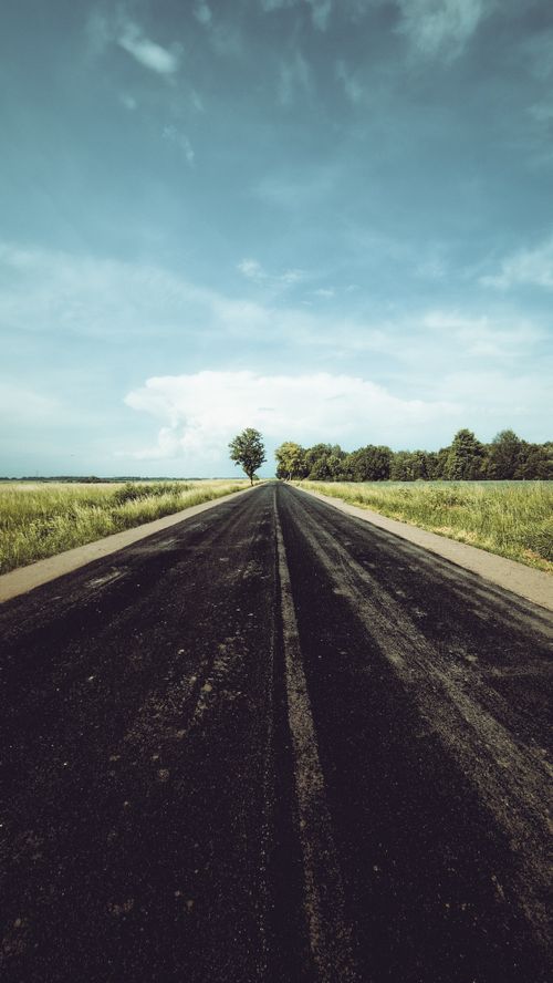 Free A straight asphalt road stretches through lush countryside fields under a blue sky, offering a serene rural perspective. Stock Photo