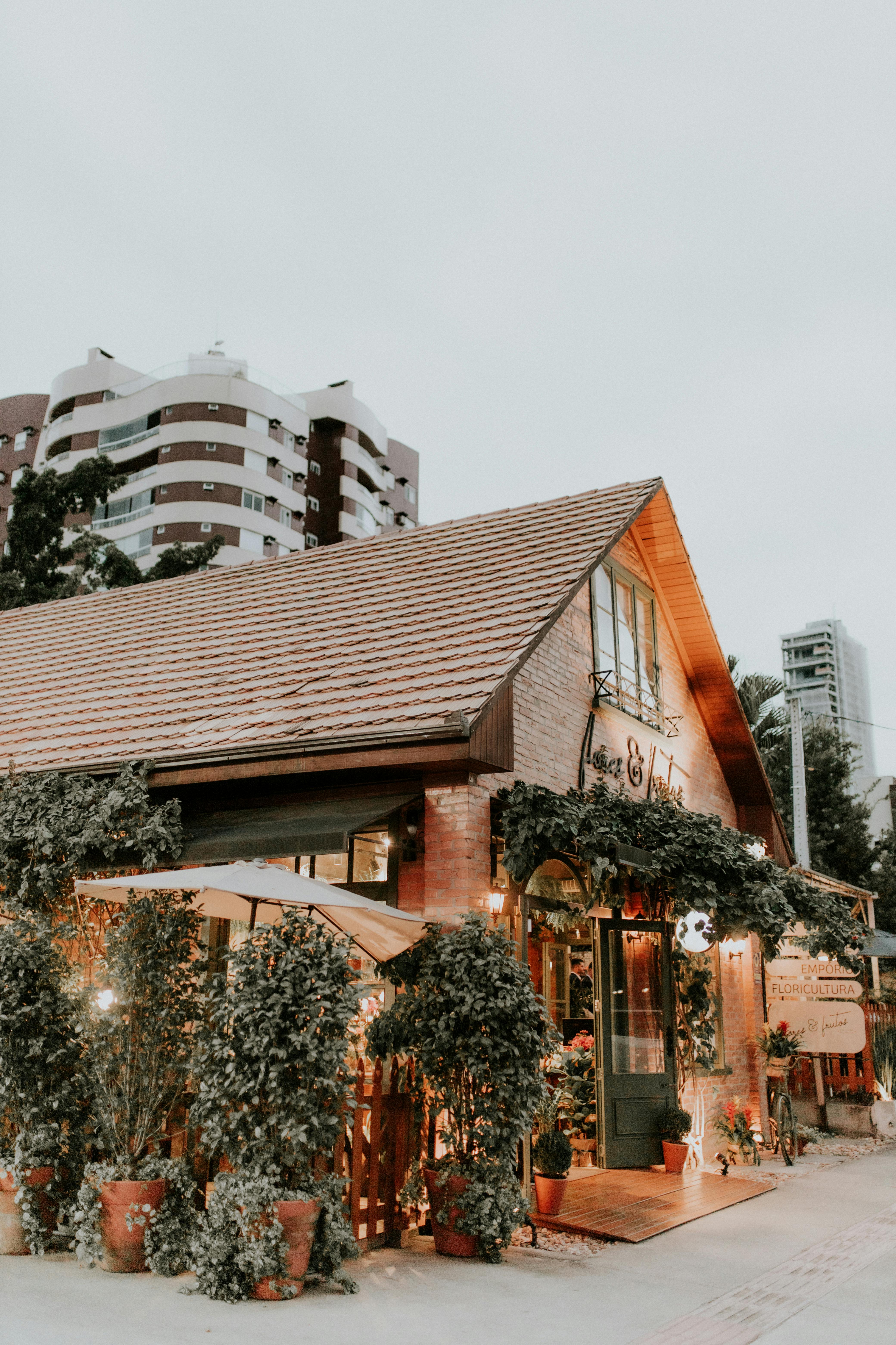 Cozy brick restaurant facade with potted trees, urban backdrop, evening ambiance.