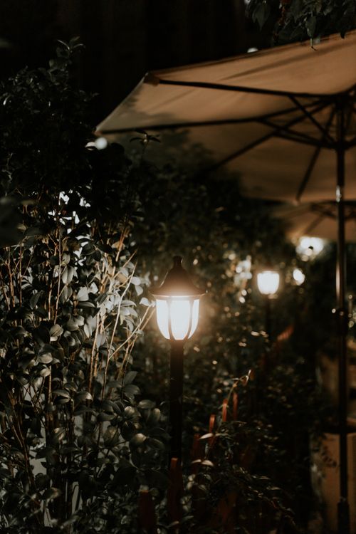 Free Illuminated garden path with lanterns, umbrellas, and shrubs creating a tranquil nighttime atmosphere. Stock Photo