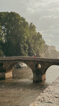A person with an umbrella crosses a vintage stone bridge over a river during a light summer rain.