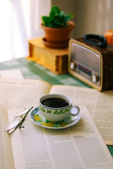 Cup of coffee on an open magazine with vintage radio and plant, creating a cozy indoor setting.