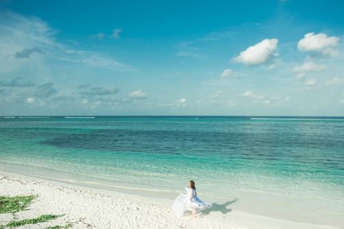 Free A woman in a flowing white dress walks along a serene tropical beach under a clear blue sky. Stock Photo