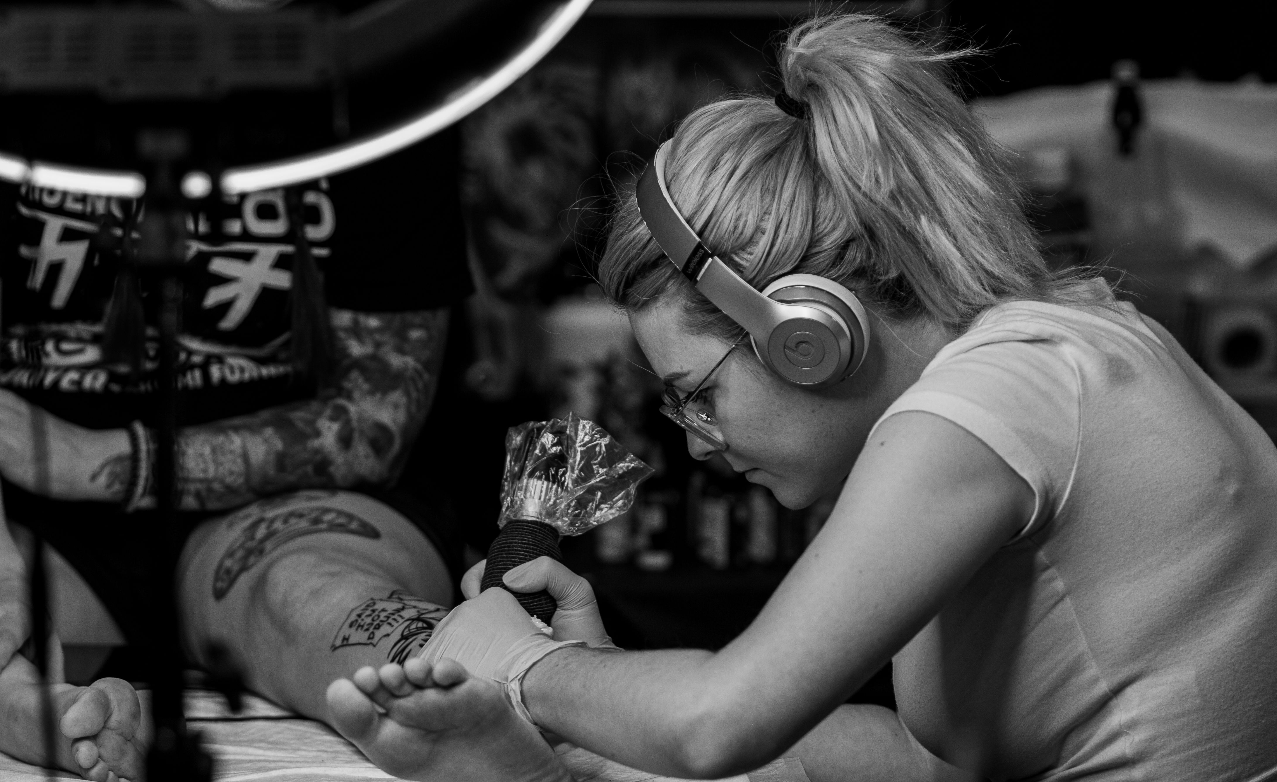 Black and white photo of a tattoo artist working on leg art in a professional studio setting.