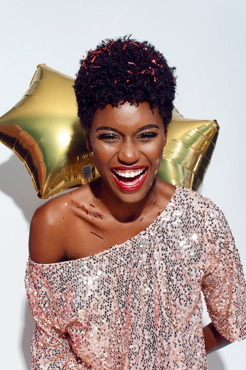 Photo of Laughing Woman with Confetti on Her Hair and Shoulders Posing In Front of White Background While Holding Golden Star Balloons Behind Her Back