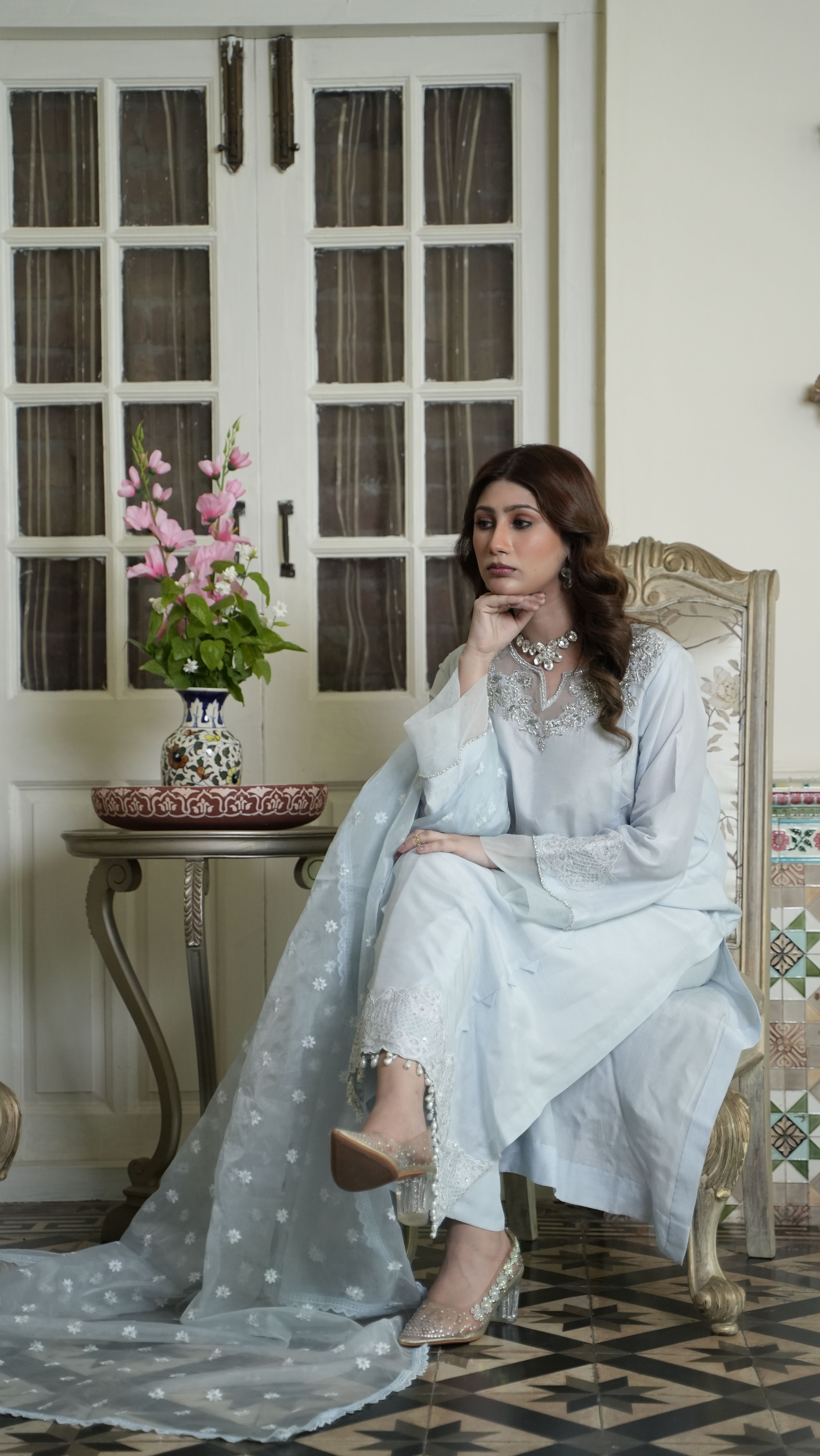 A woman in an elegant white dress and shawl sitting indoors, showcasing traditional fashion and elegance.