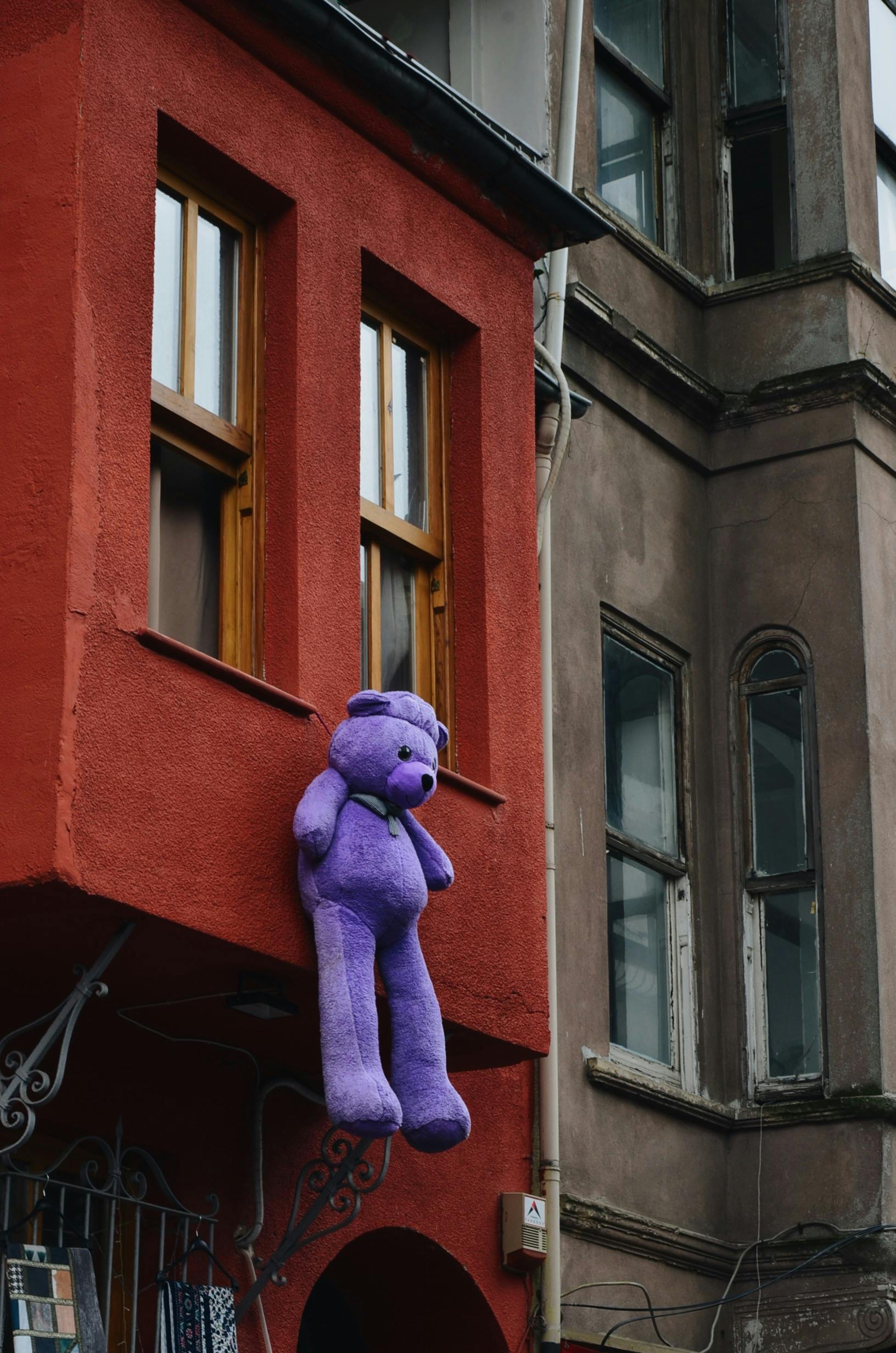 Free A purple teddy bear hangs from a red urban building window, creating a whimsical city scene. Stock Photo