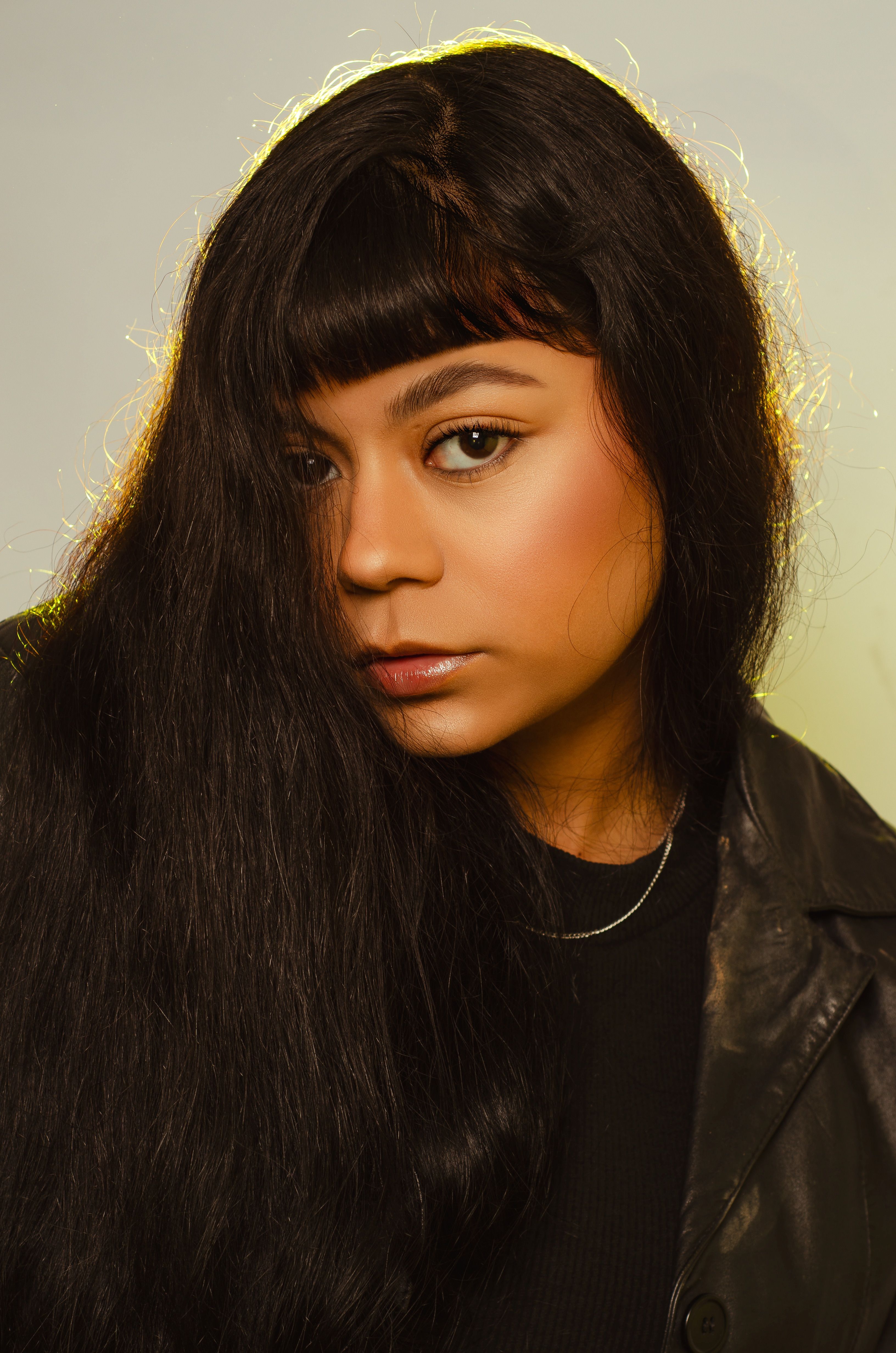 Close-up portrait of a woman with long black hair, wearing a leather jacket, shot in a studio setting.