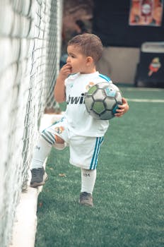 A child in sportswear holding a soccer ball on a football pitch, exhibiting innocence and curiosity.