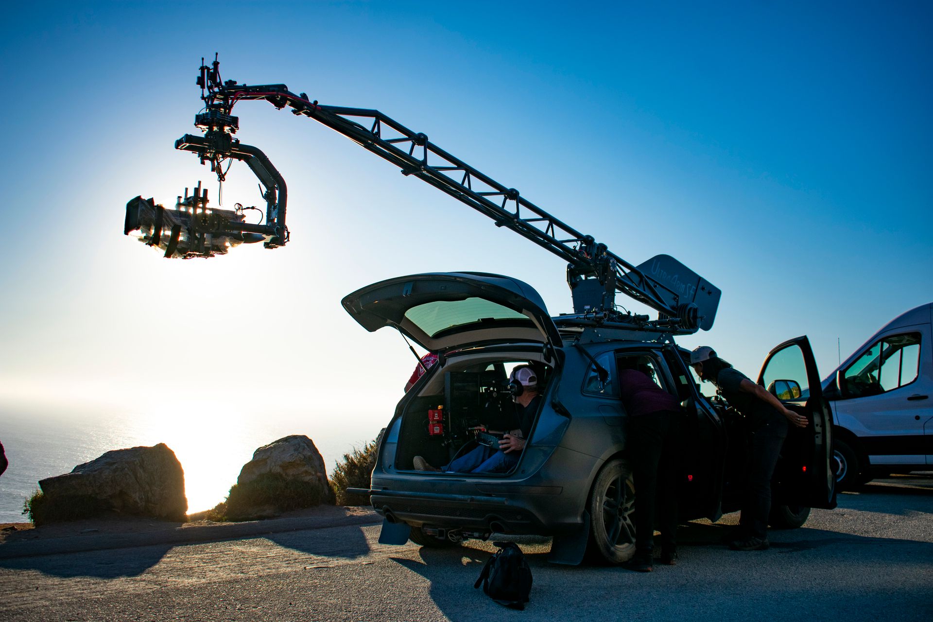 A film crew operates a camera crane mounted on a vehicle at a cliffside location during sunset.
