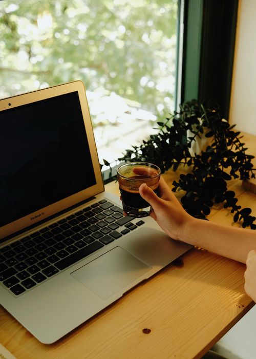 Free A hand holding coffee next to laptop on wooden desk by window with potted plant in İstanbul. Stock Photo