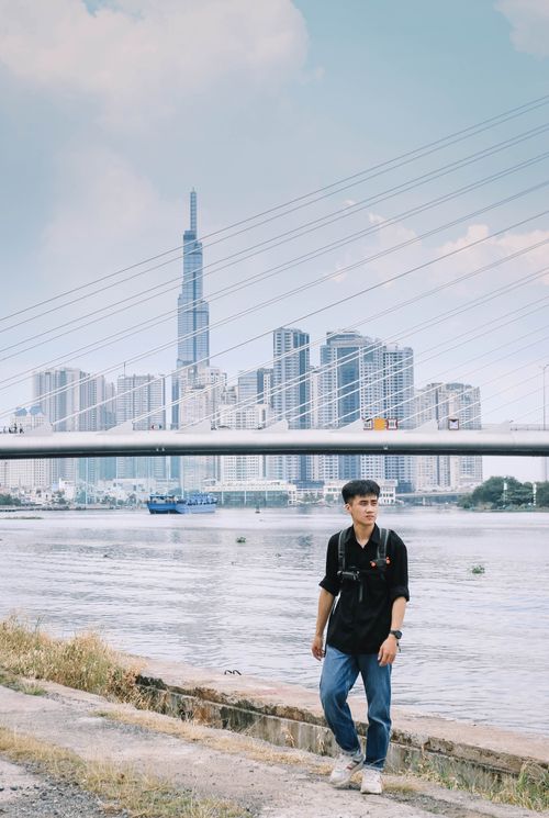 Free Young man walking by river with Ho Chi Minh City skyline and bridge in background, showcasing modern urban cityscape. Stock Photo