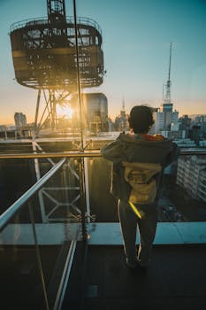 A person enjoys a sunset view of the city skyline from a rooftop with glass railings, creating a modern urban scene.