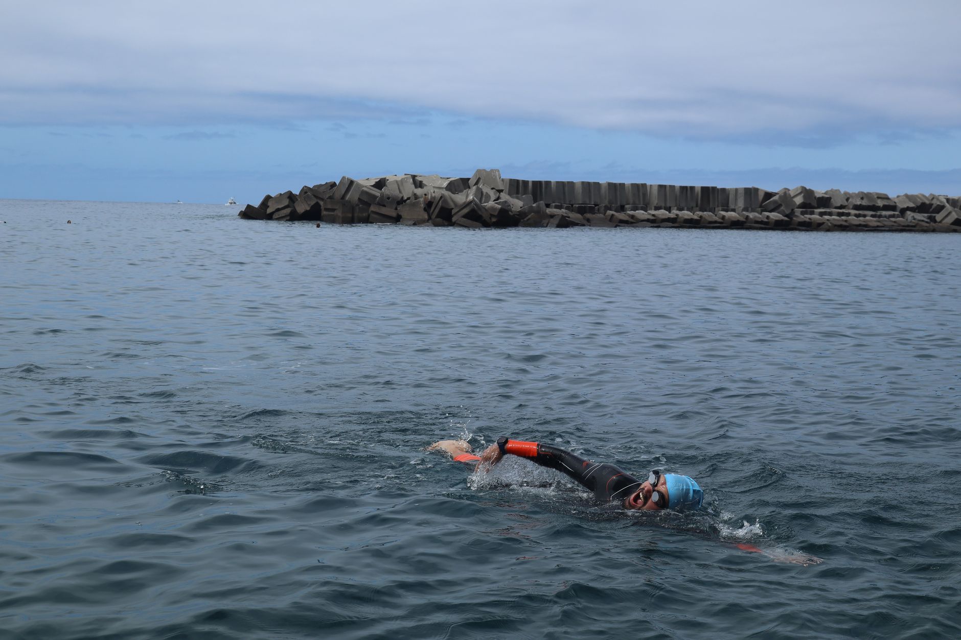 A swimmer in a wetsuit, blue swim hat and orange tow float crossing calm grey sea near a breakwater on an overcast day.