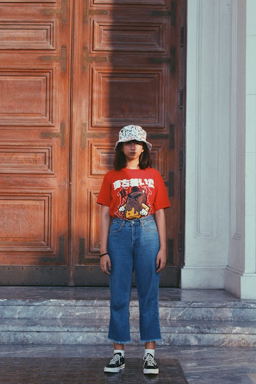 Photo of Teenage Girl in Bucket Hat, Red T-shirt, and Blue Jeans Standing In Front of Brown Wooden Door