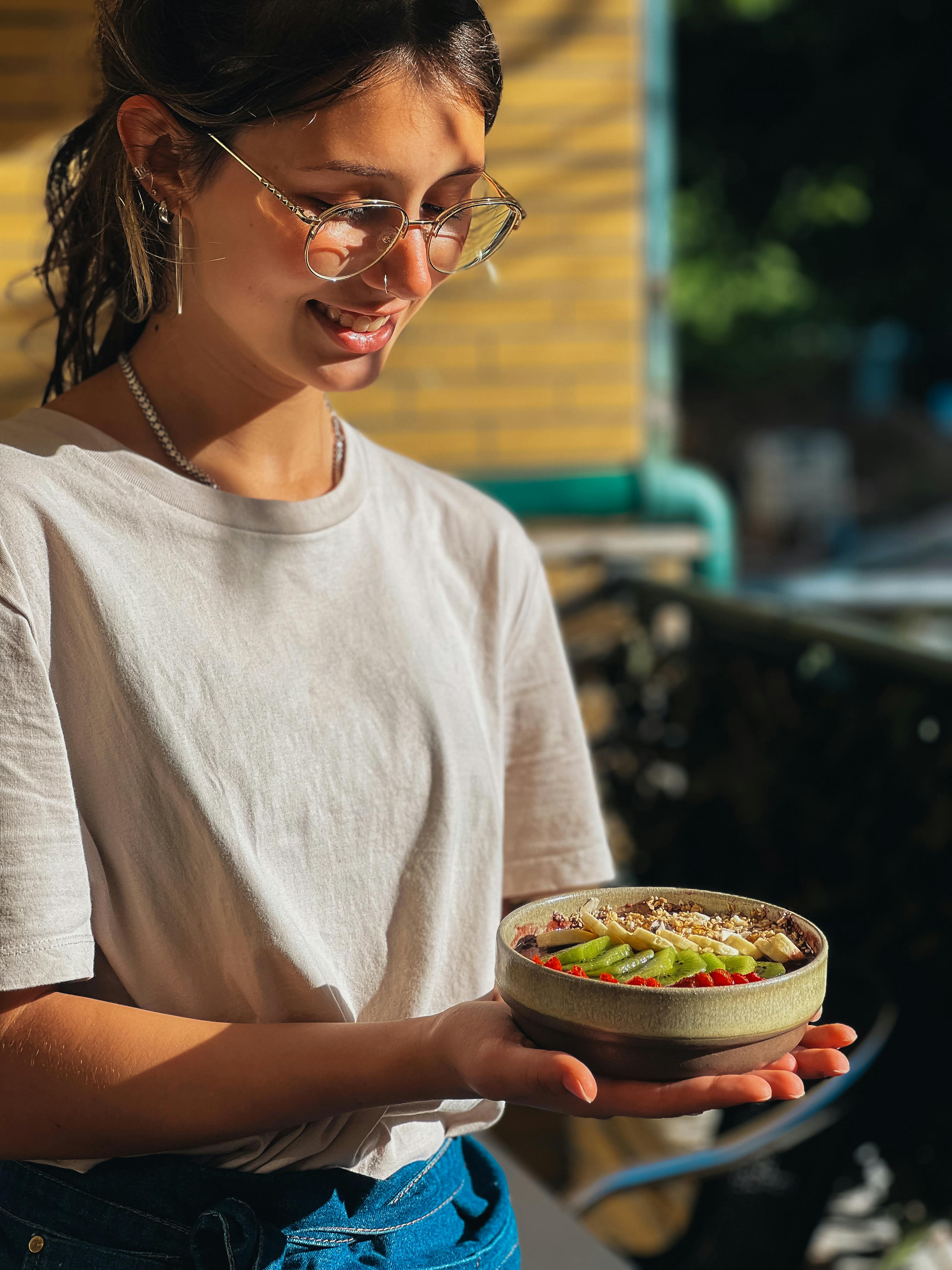 Smiling woman with eyeglasses holds a nutritious bowl outdoors in sunlight.