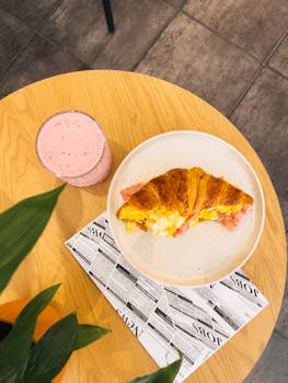 Enjoy a delicious breakfast featuring a croissant sandwich and smoothie set on a wooden table.