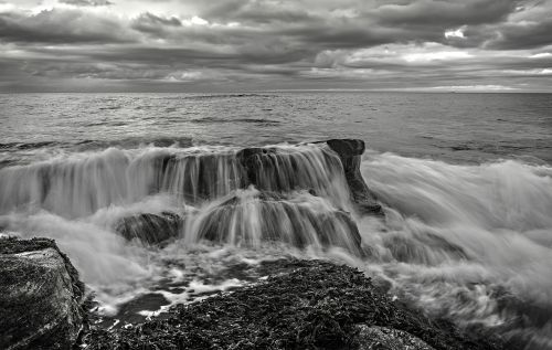 Free Dynamic black and white seascape with waves crashing over rocky shoreline under a moody sky. Stock Photo