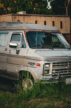 Classic silver van in tall grass with building backdrop under warm sunlight.